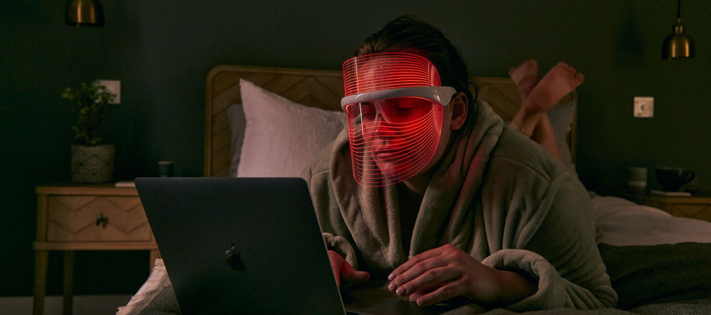 Woman laid on bed wearing MAGNITONE GetLit LED Light Mask while using laptop
